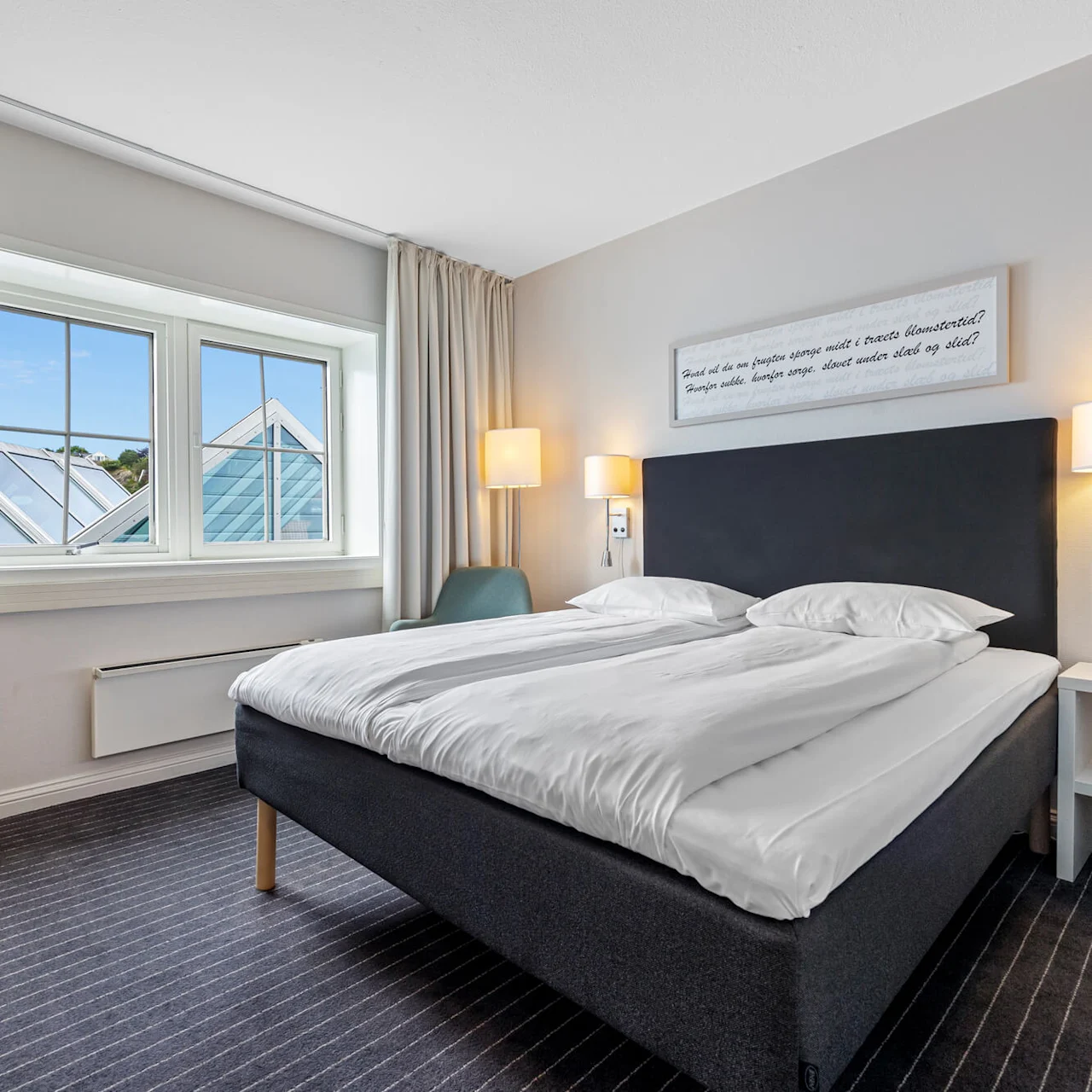A tidy hotel room with an unmade bed, facing a window with geometric roof details; a framed text is above the bed.