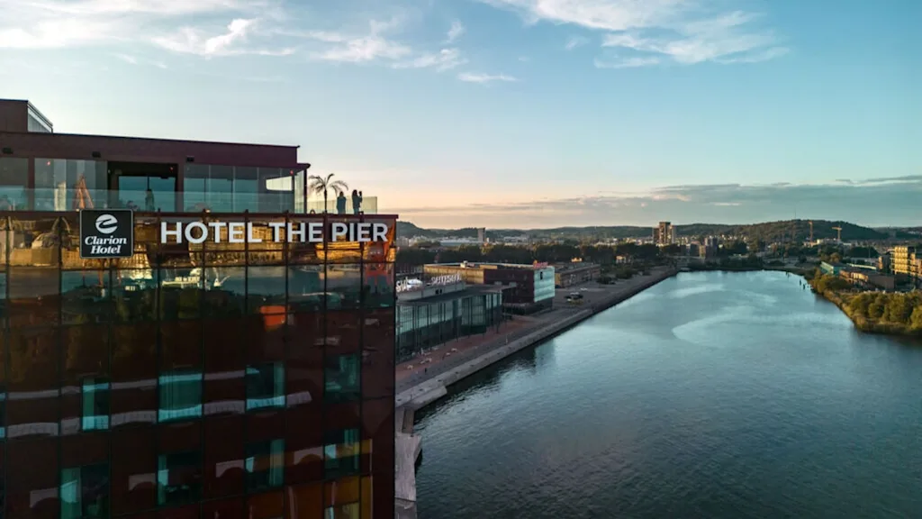 A waterfront hotel building labeled "Clarion Hotel THE PIER" stands beside a calm river, framed by a sunset-lit cityscape extending into the distance.