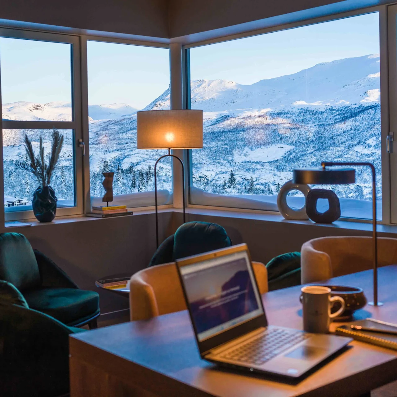 Laptop on a wooden desk, displaying a blurred screen, with notebooks and a mug. Surrounded by chairs, contemporary art pieces, and large windows showcasing a snowy mountain landscape.