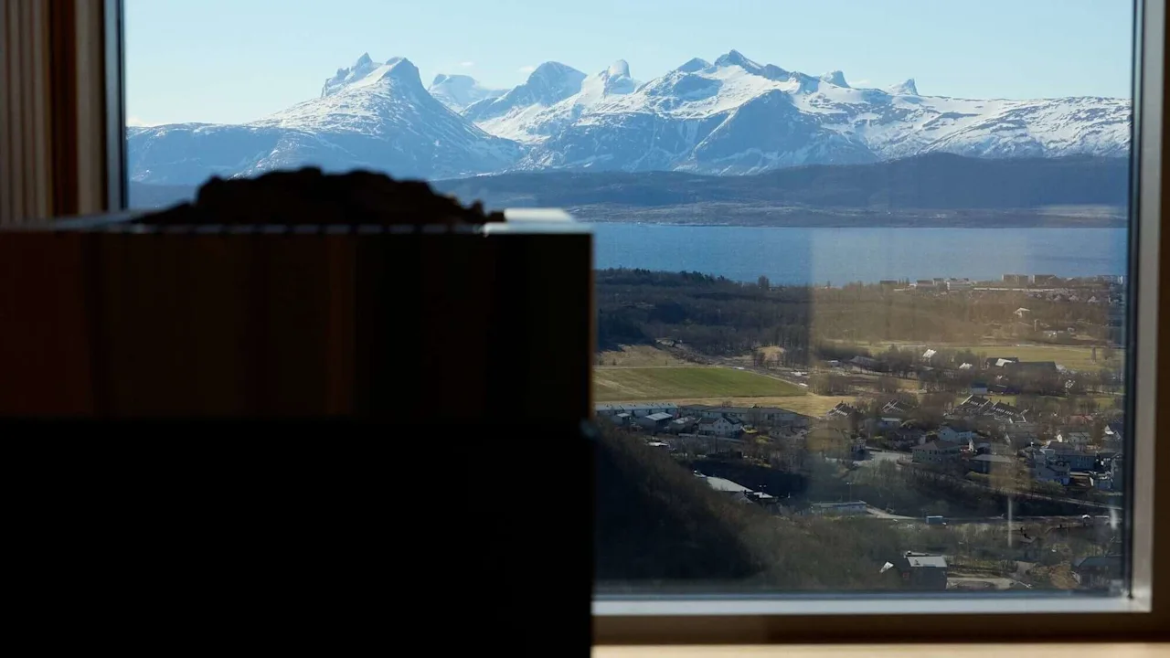 En bastuugn är i silhuett i förgrunden, med snötäckta berg synliga genom ett stort fönster, med utsikt över en stad under en klarblå himmel.