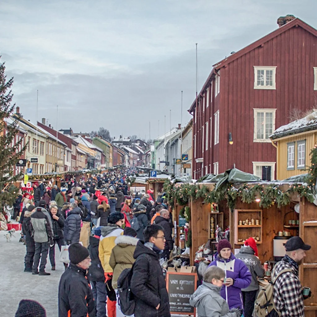 A lively outdoor market scene with many people browsing stalls and walking on a snowy street, featuring a large Christmas tree.