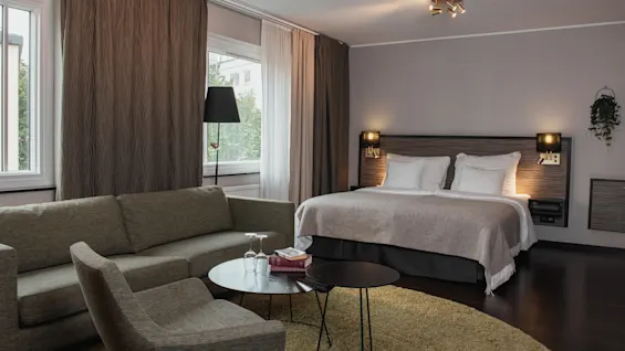 Viihtyisä hotellihuone, jossa on suuri sänky, mukava sohva ja nojatuoli, lämpimällä valaistuksella ja luonnonvalolla ikkunoista.