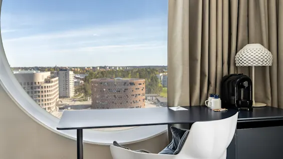 Et moderne hotelværelses skrivebord med et rundt vindue med udsigt over en by, med en kaffemaskine og en stilfuld lampe.