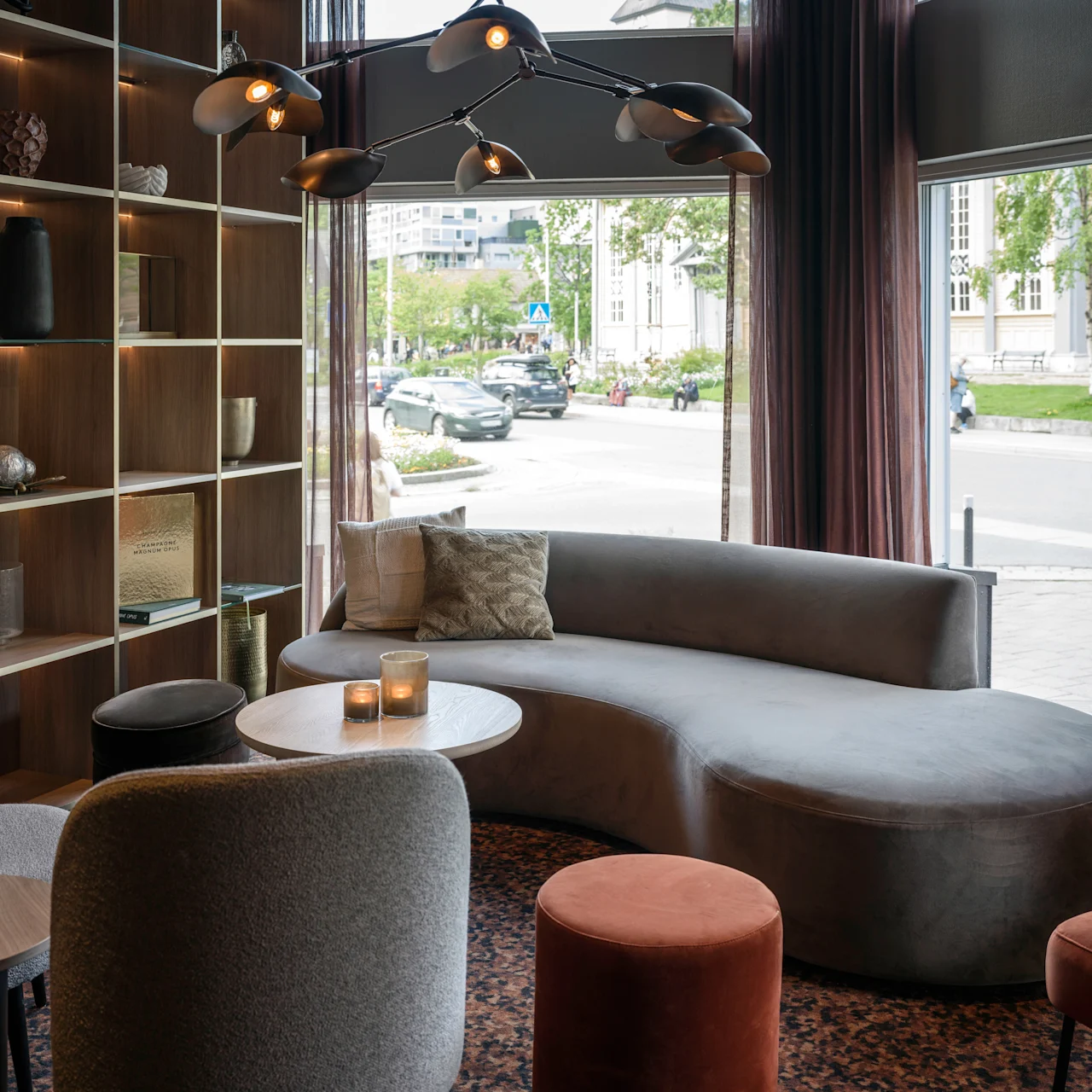 A curved gray sofa sits in a modern lounge, surrounded by chairs and a wooden shelf with decor items. Large windows reveal a street with trees and parked cars outside.