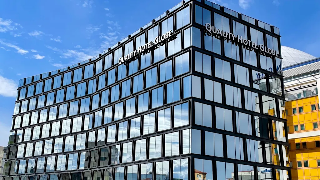 A modern glass building displays "QUALITY HOTEL GLOBE" on the facade, reflecting sunny blue skies. It stands against a city backdrop with nearby yellow and white structures.