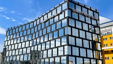En modern glasbyggnad visar "QUALITY HOTEL GLOBE" på fasaden och speglar en solig blå himmel. Den står mot en stadsmiljö med närliggande gula och vita byggnader.