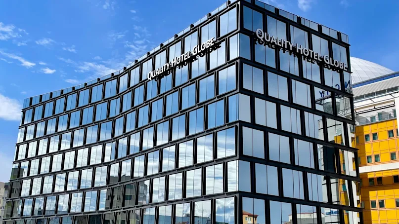 A modern glass building displays "QUALITY HOTEL GLOBE" on the facade, reflecting sunny blue skies. It stands against a city backdrop with nearby yellow and white structures.