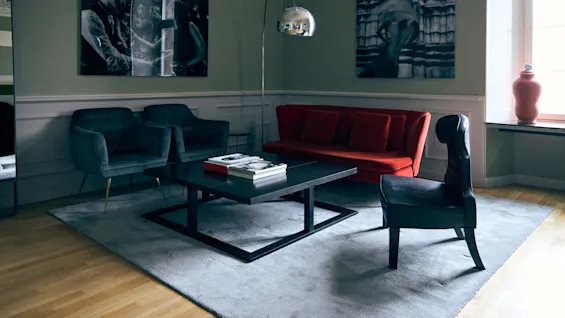 A modern living room with a black coffee table on a gray rug, surrounded by two blue chairs, a red sofa, and a black chair. Black-and-white photos decorate the walls.