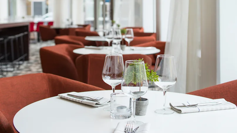 Round white table set with empty wine glasses, potted plant, and napkin-wrapped cutlery in a modern restaurant with red armchairs and large windows.