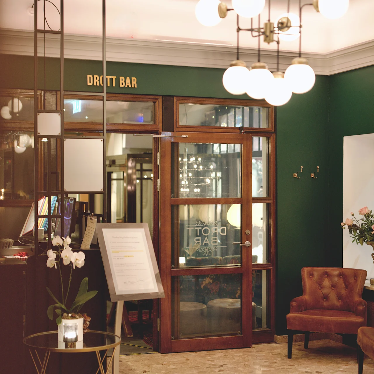 En lysekrone hænger i en hotellobby med marmorgulv, brune stole og orkidéer i potter. Et skilt siger "DROTT BAR" over en dør af glas.