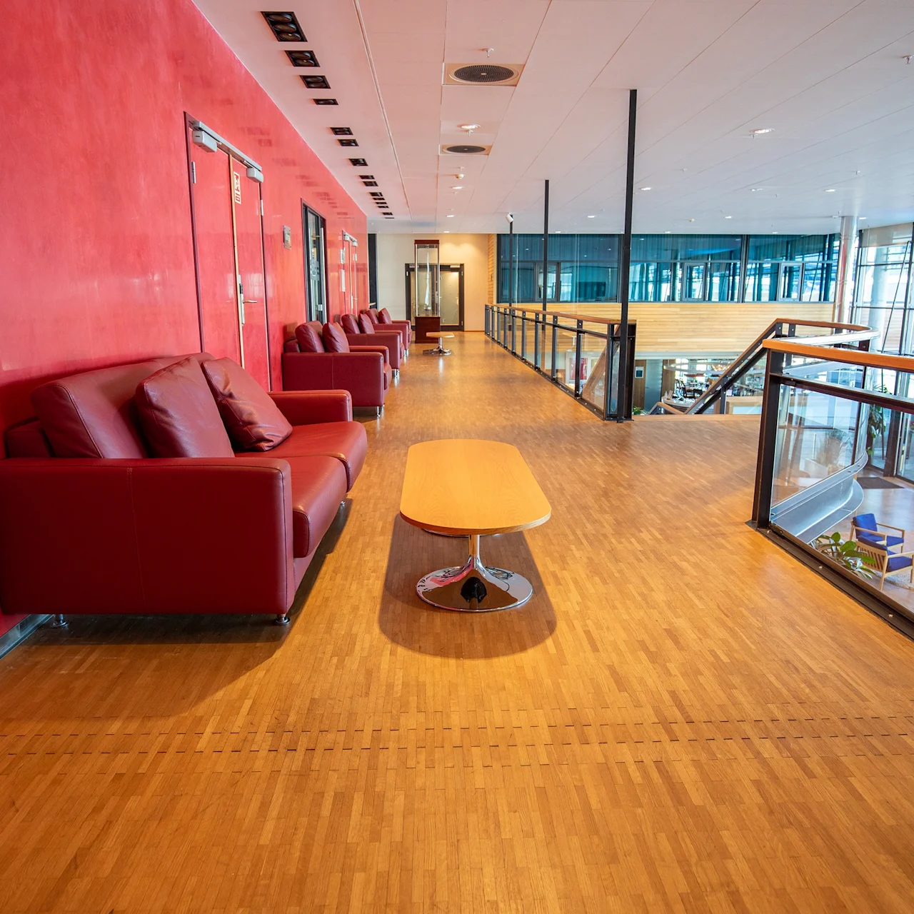 Røde sofaer står langs en gang med tregulv ved siden av et glassrekkverk, med utsikt over et åpent atrium med ekstra sitteplasser.