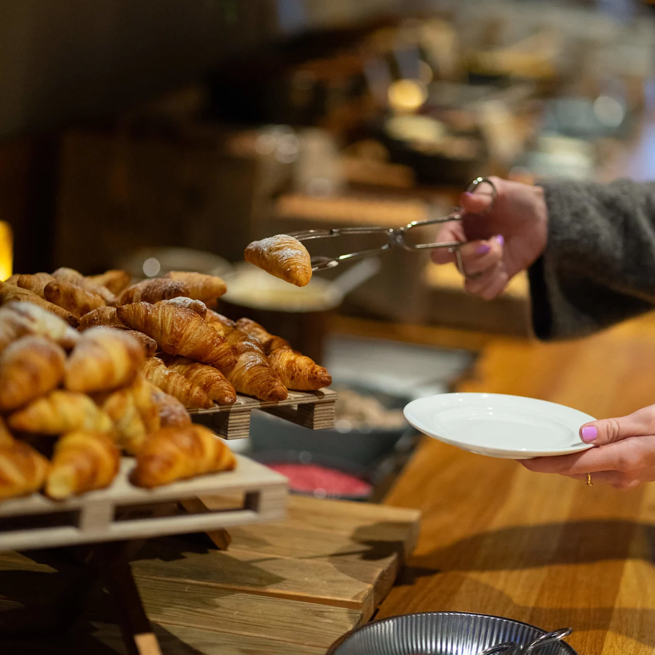 Croissant ligger på en trestativ; en person bruker tang til å legge en på en hvit plate, i en buffésetting.