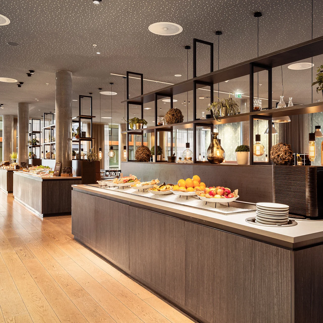 Buffet tables displaying fruits and various dishes are arranged in a modern dining area with wooden floors and decorative lighting.