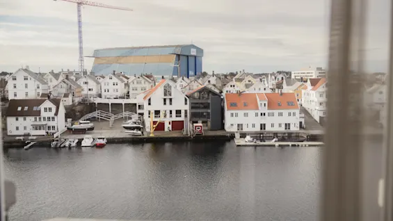 Vinduesudsigt over en charmerende kystby med hvide og røde taghuse langs en havn fyldt med både, under overskyet himmel.