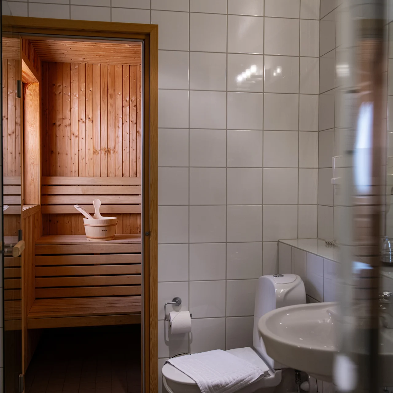 A sauna with wooden walls and a bench is visible through a doorway, connected to a tiled bathroom with a sink, toilet, and a mirror reflecting the scene.