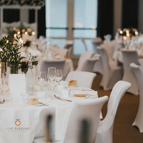 Elegant dekket rundt bryllupsbord med hvite duker, stoltrekk, vinglass, levende lys og oppsatser med hvite roser.