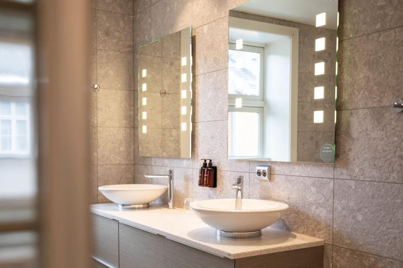 To moderne hvite vasker med kromkraner står på en beige benkeplate under opplyste speil. Grå flislagte vegger og et vindu gir et lyst, elegant baderomsmiljø.
