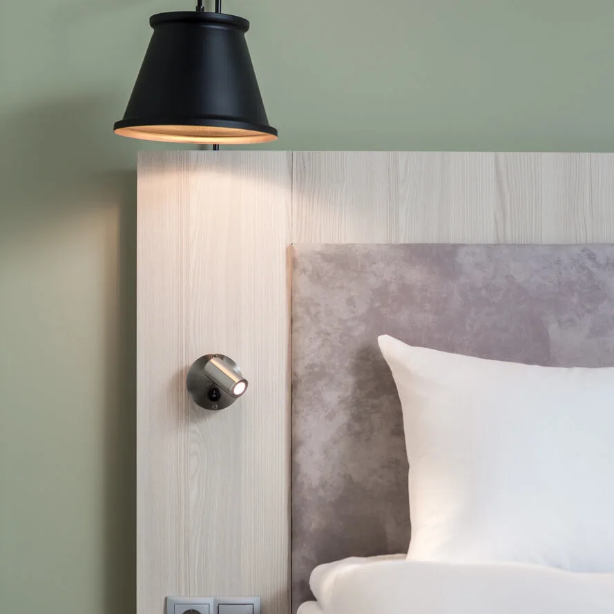A black wall lamp illuminates a beige headboard beside a neatly made bed with white pillows, set in a room with light green walls and an electrical outlet nearby.