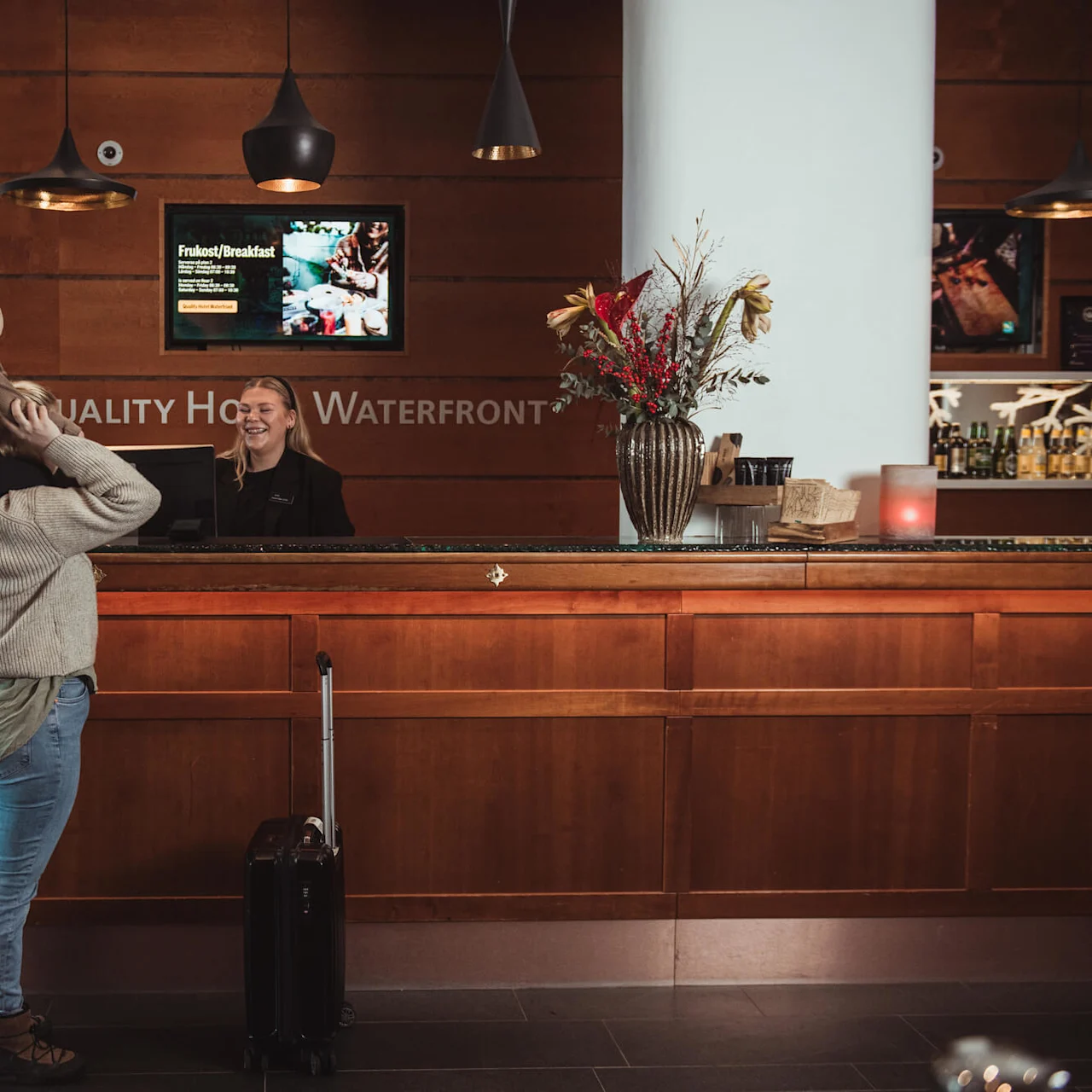 A person holds a child on their shoulders, standing at a hotel reception desk. A receptionist smiles behind the counter. The wall displays "QUALITY HOTEL WATERFRONT". A suitcase and flowers are nearby. Text: "Frukost/Breakfast".
