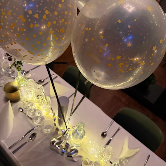 Festligt dukat middagsbord med glittrande ljusslingor, elegant porslin och genomskinliga ballonger fyllda med guldkonfetti.