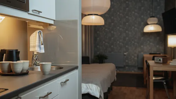 Kitchen countertop with a sink and coffee maker holds three white cups; illuminated, modern interior includes a bed, wooden table, and hanging lamps in a cozy, softly lit room.  