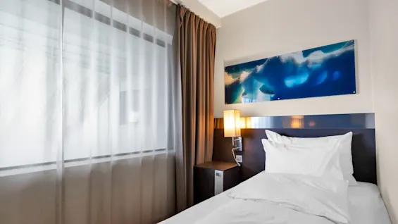 A neatly made single bed with white linens is beside a wall lamp and nightstand; sheer curtains cover a large window, and abstract blue artwork adorns the wall.
