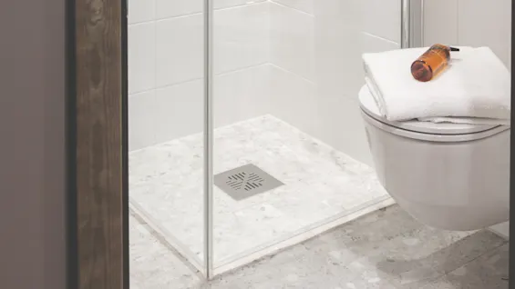 Dusjkabin av glass; har en montert dusjhode, gulvavløp og tilstøtende toalett med foldede håndklær og flaske på toppen; hvite flislagte vegger i et moderne bad.
