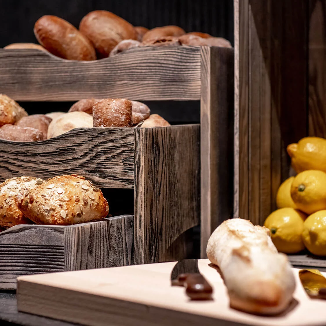 Brød ruller er stablet i trekasser. En skjærebrett har en baguette og kniv. Lyse gule sitroner fyller en kasse i bakgrunnen.
