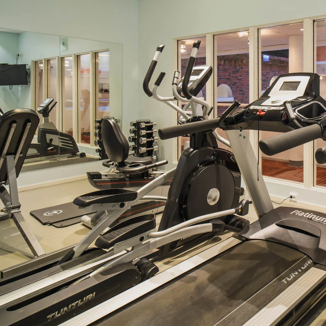Treadmills and exercise machines are arranged in a gym with mirrors and windows. Two treadmills display "TUNTURI" and "Platinum" on their fronts.