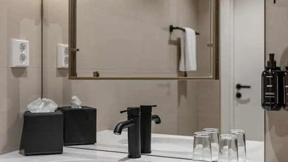 Et moderne badeværelse med hvid vask, sort hane og stort spejl. Tissuebokse, glas og sæbe samt body lotion på bordet.