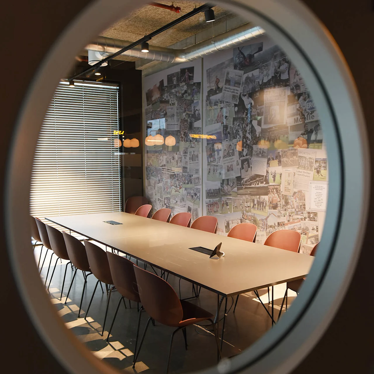 Et langt konferencetable omringet af stole i et rum med store vinduer og en væg dækket af collagebilleder, set gennem et cirkulært glasvindue.
