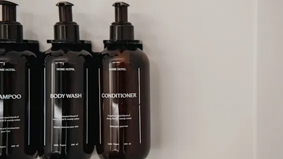 Tre mørke flasker med pumper mærket SHAMPOO, BODY WASH og CONDITIONER, monteret på en væg.