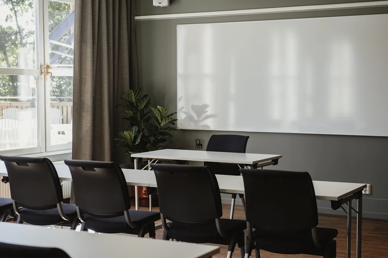 Ett ljust mötesrum med rader av bord och stolar vända mot en stor whiteboard. Ett fönster med gardiner finns till vänster.