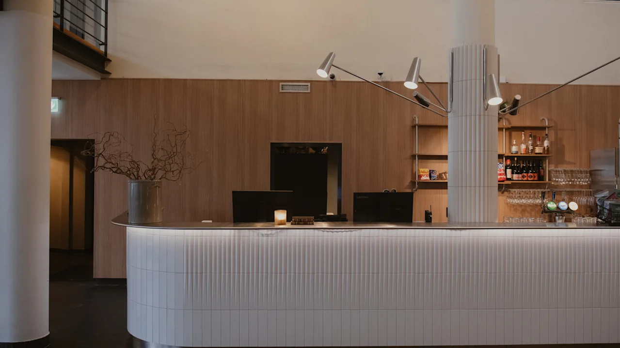 En moderne resepsjonsdisk med buet design står i en varmt opplyst lobby. Bak den, trehyller viser flasker, og en elegant lysarmatur henger over.