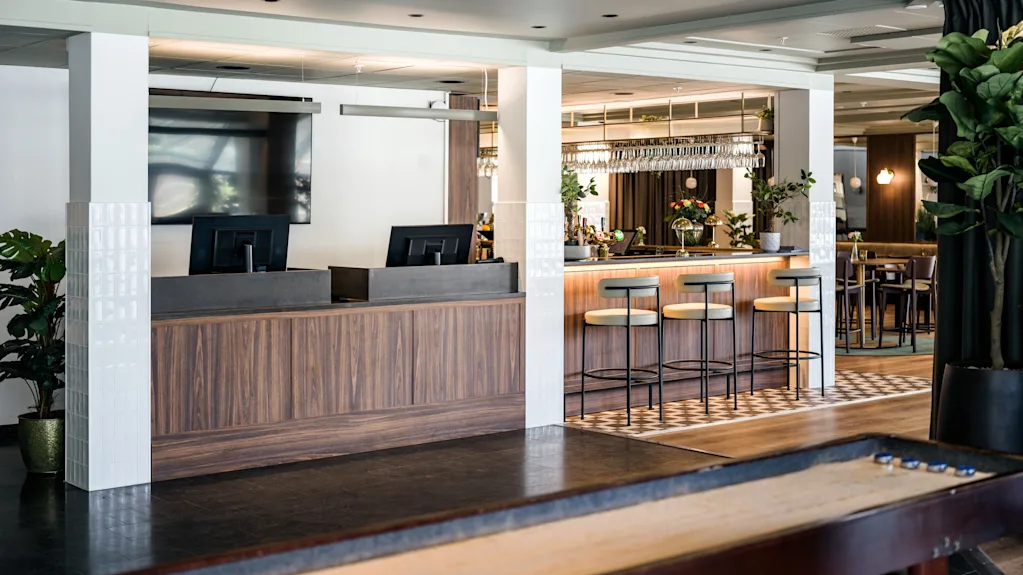 Resepsjonsdisken står tom ved siden av en moderne bar med høye krakker, omgitt av inneplanter og stemningsfull belysning. Et shuffleboard-bord opptar forgrunnen.