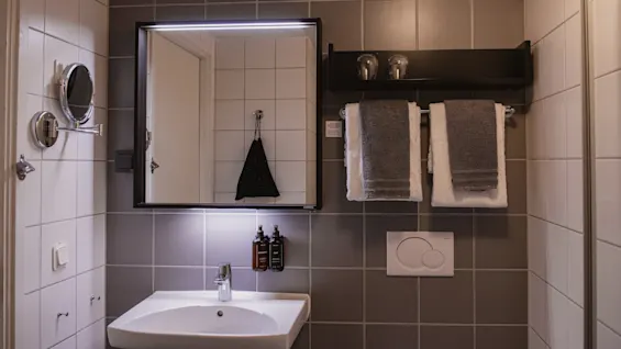 Ett badrum har ett handfat med spegel ovanför och tvålpumpar. Handdukar hänger på en ställning bredvid en toalettspolning mot grå kaklade väggar.