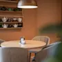 A round wooden table with salt and pepper shakers and a candle stands surrounded by gray chairs. Shelves with various teapots hang on a brown wall under a metal pendant light.