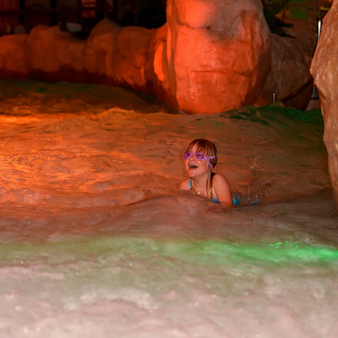 A child wearing goggles is swimming in shallow, illuminated water surrounded by artificial rock formations in a dimly lit indoor environment.