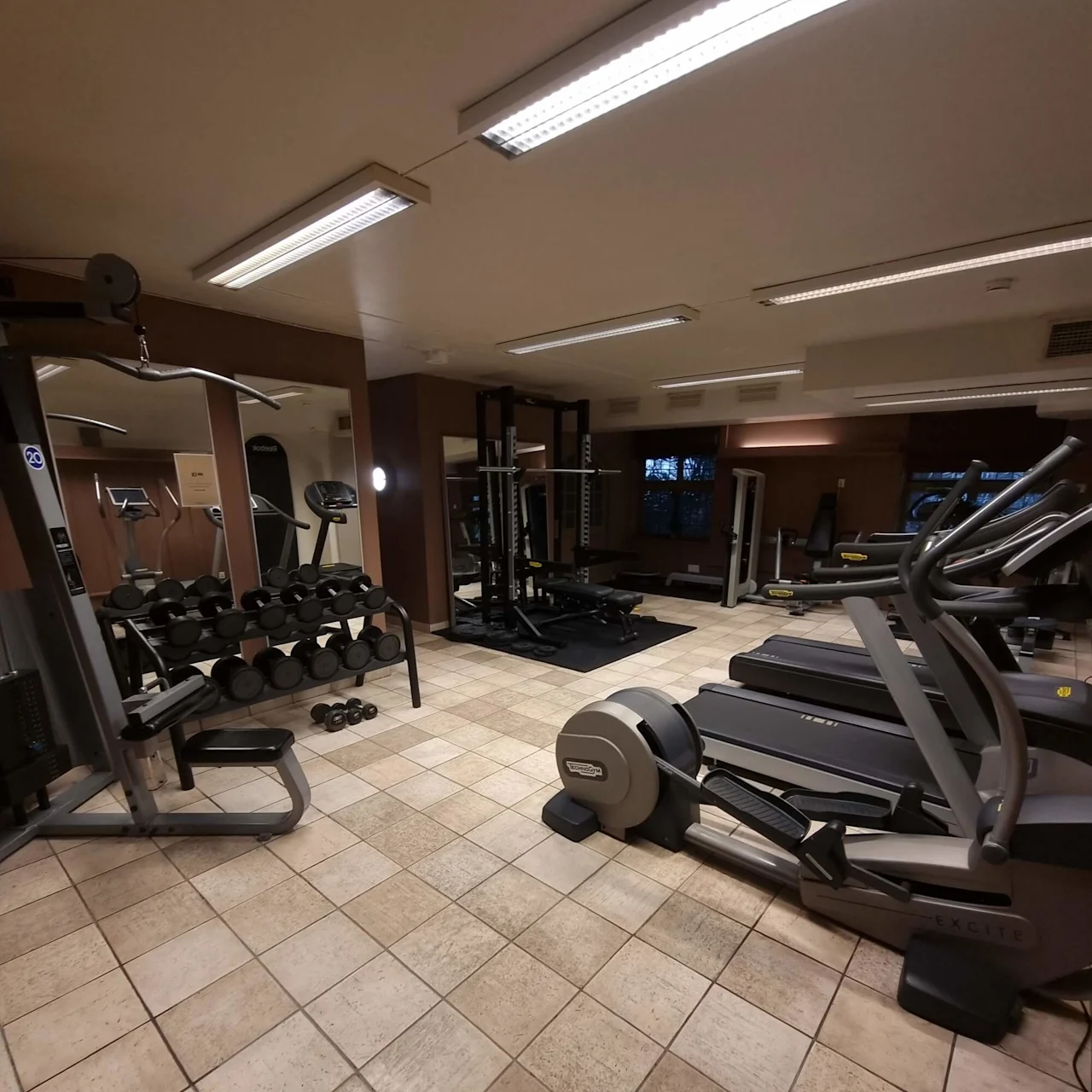 Träningsutrustning i ett tomt gym med löpband, vikter och maskiner på kaklat golv.
