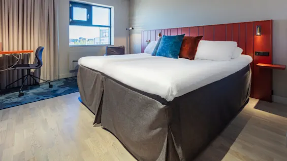 A neatly made bed centered in a well-lit, modern hotel room with curtains, a desk, chair, and an open window showing a cityscape.