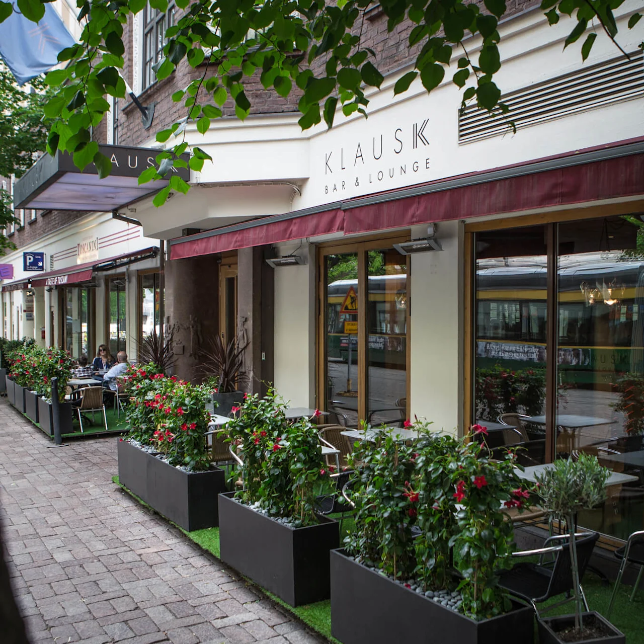 Baari ja lounge nimeltään "KlausK", jossa on ulkopöytiä ja tuoleja suurten istutusten vieressä. Se sijaitsee puiden reunustamalla kadulla.