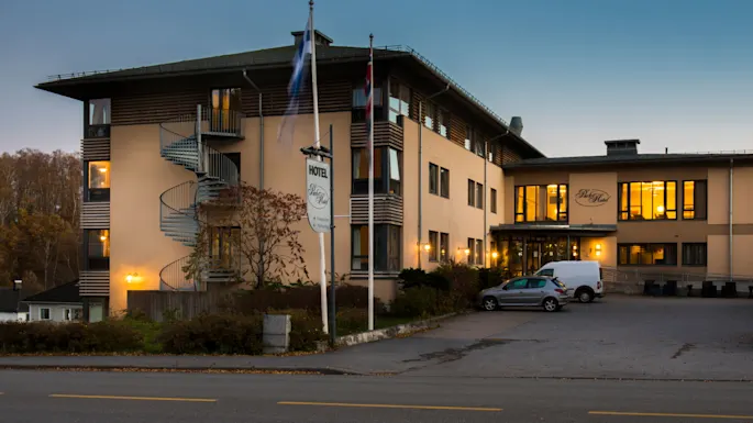 A hotel at dusk with illuminated windows, parked cars, and flapping flags in front.