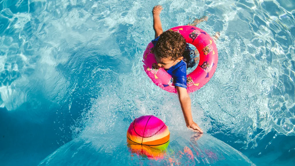 A child in a pink swim ring reaches for a colorful ball in a bright blue swimming pool with splashing water.