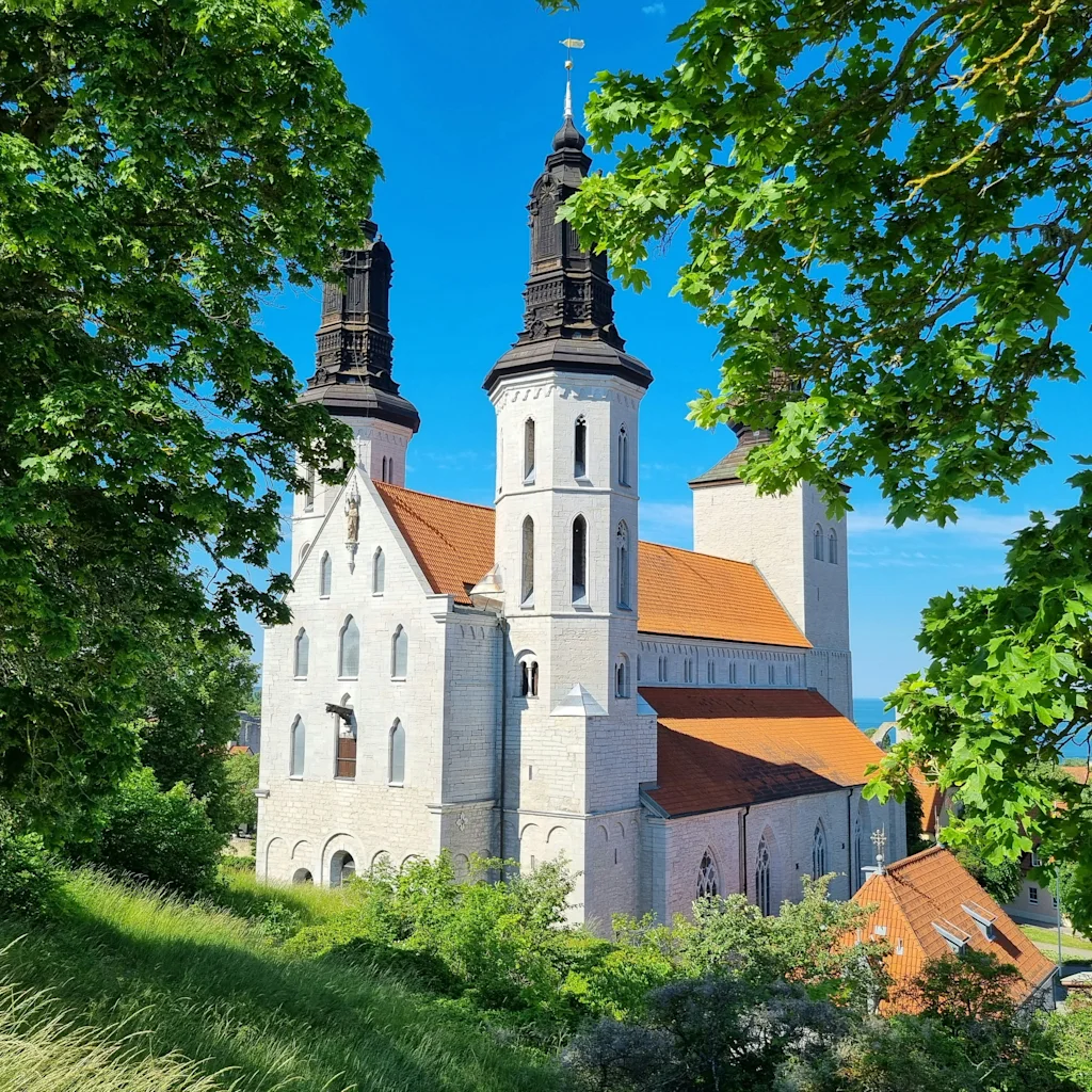Suuri, vaalea rakennus, jossa on kaksi korkeaa tornia ja oranssit katot, rehevien vihreiden puiden kehystämänä kirkkaan sinisen taivaan alla.