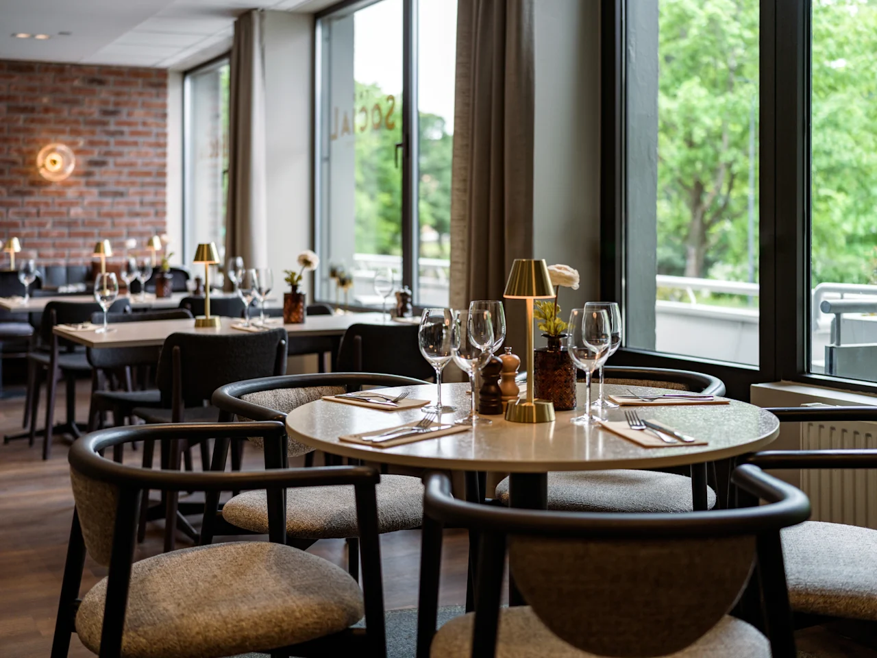 Modernt matbord med vinglas, bestick och en liten lampa i en restaurang med tegelväggar och stora fönster som vetter mot gröna träd.