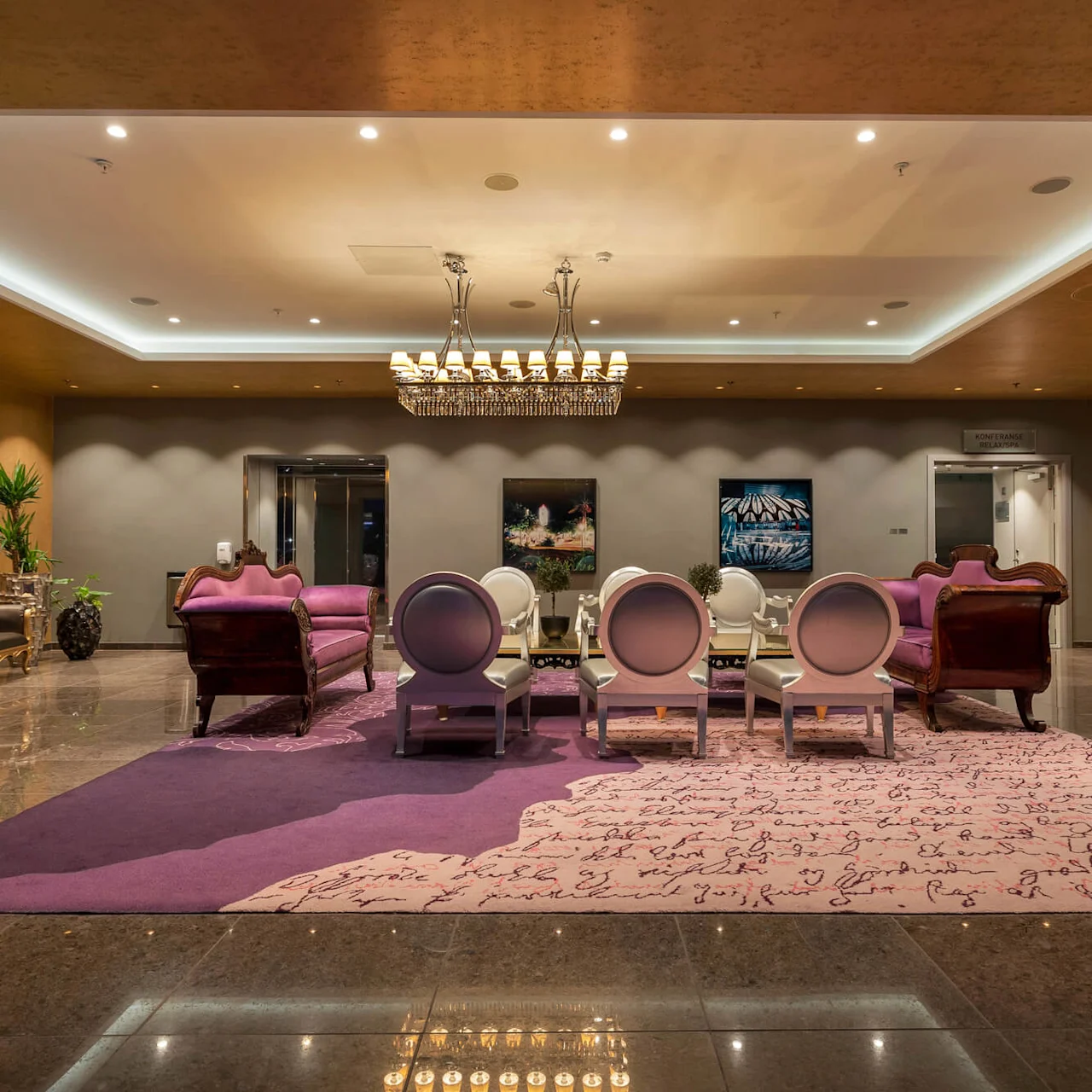 Luksuriøs lounge med lilla sofaer og runde stole omkring et glasbord. Elegant lysekrone over, foran kunstprydede vægge. Tekst: "KONFERANSE RESEPSJON".