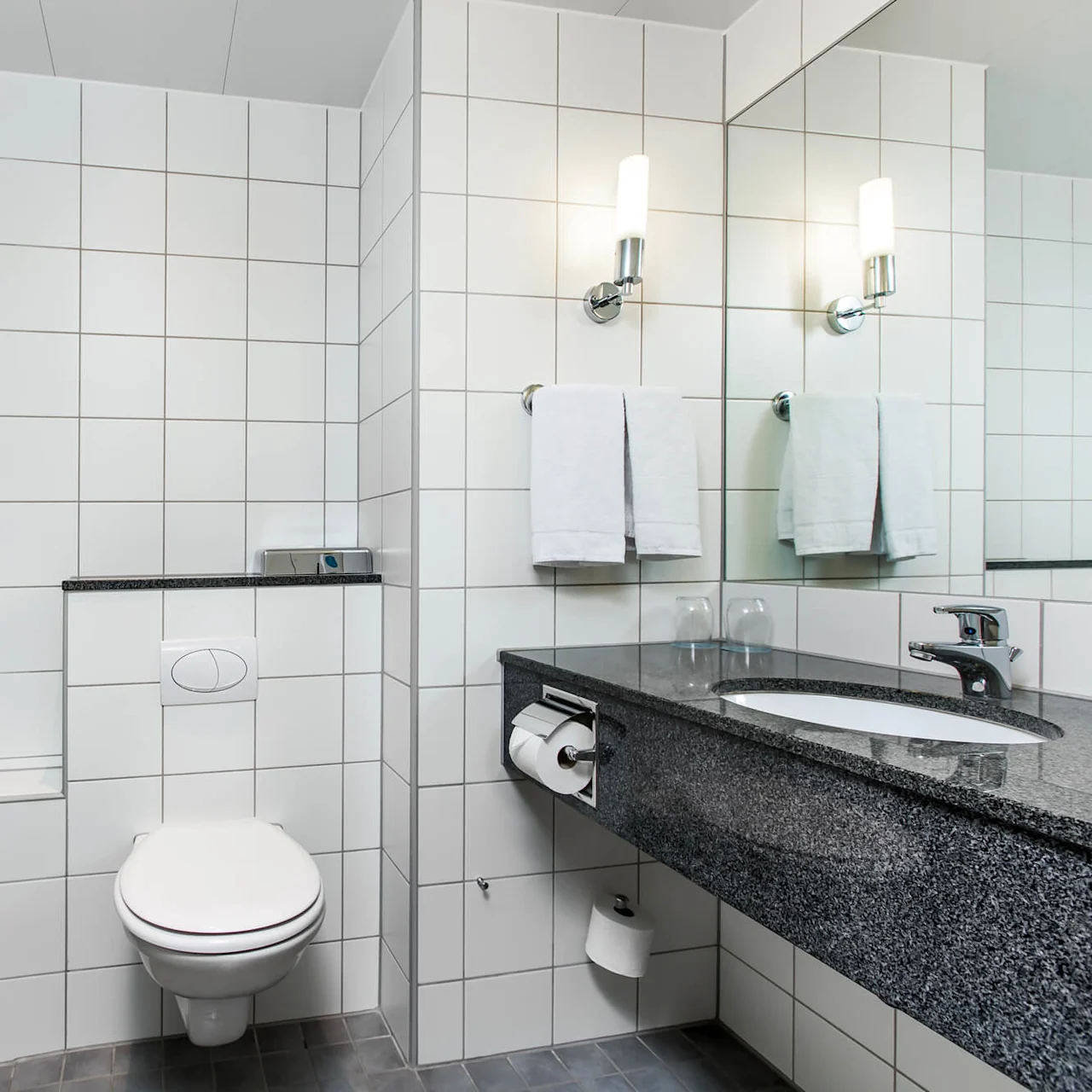 A bathroom with a bathtub and shower combo, toilet, and a granite sink counter adorned with towels and glasses. White tiles cover walls and floor, creating a clean, modern look.