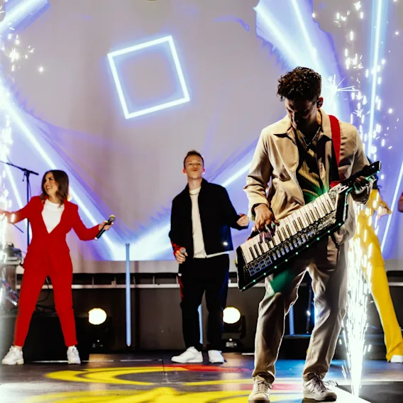 Live band på scenen med sangere, gitarist og en person foran bildet som spiller keytar, med gnister på scenen