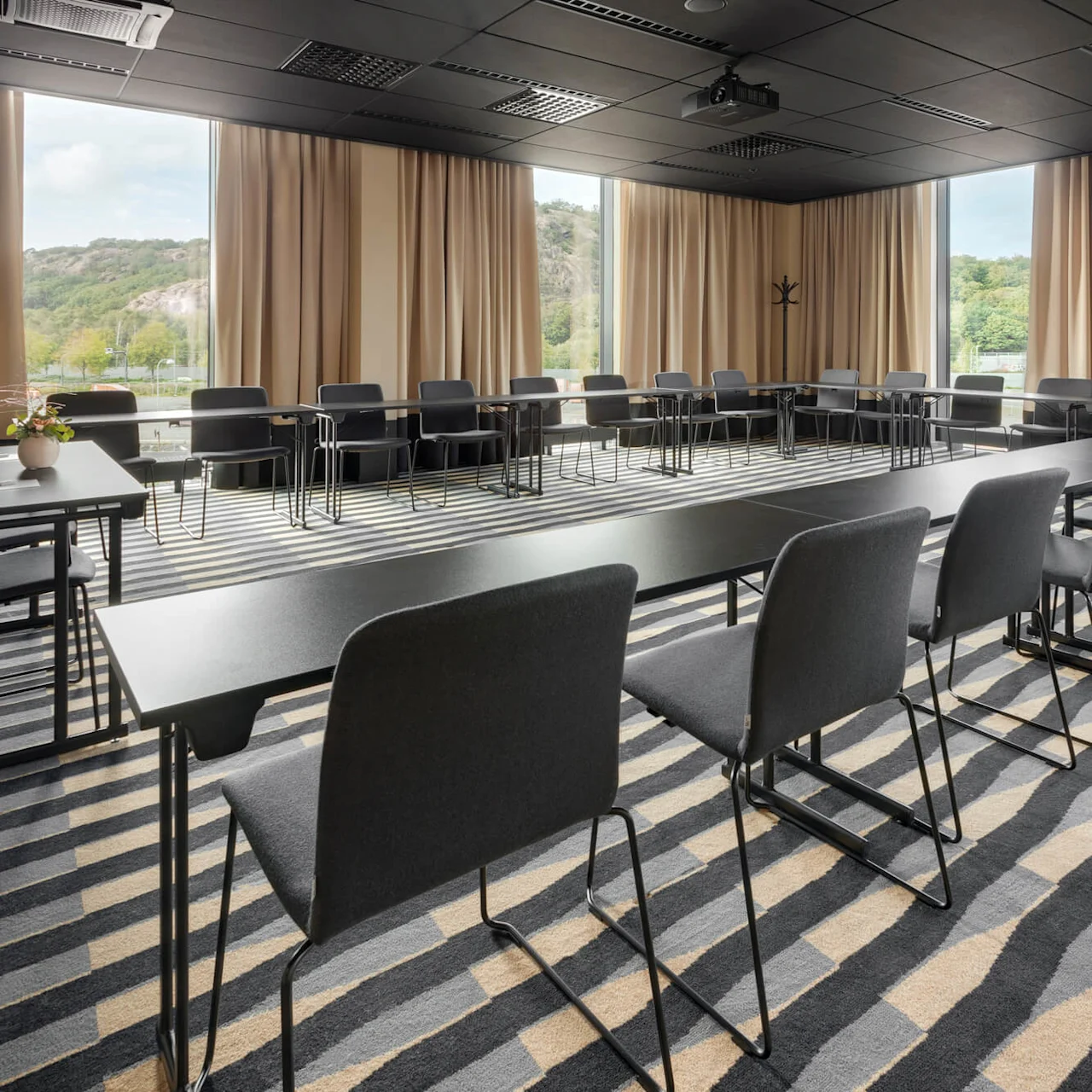 Konferensbord bildar en U-form, omgivna av grå stolar, i ett mötesrum med randig matta och stora fönster som visar ett grönt landskap utanför. Beiga gardiner ramar in fönstren.