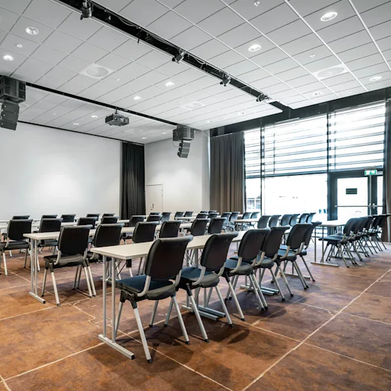 Rader av tomme stoler og bord arrangert i et moderne konferanserom, med en stor skjerm, brun teppe og høye vinduer med gardiner.